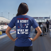 A woman stands on a racetrack from behind wearing a royal blue t-shirt with a large white and red graphic text that reads 'IT IS NOT JUST A HOBBY IT IS A WAY OF LIFE' with small red and white drag racing illustrations.