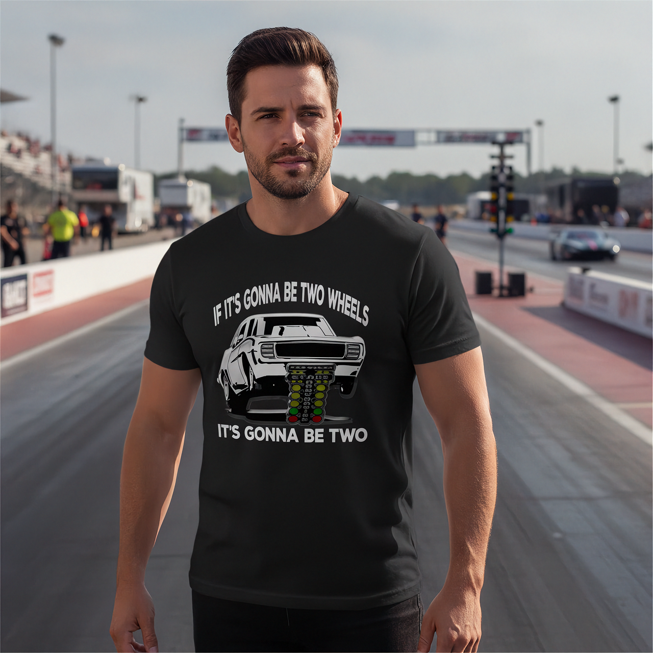A man standing on a racetrack wearing a black graphic t-shirt with a white car illustration and the text 'IF IT'S GONNA BE TWO WHEELS IT'S GONNA BE TWO'.