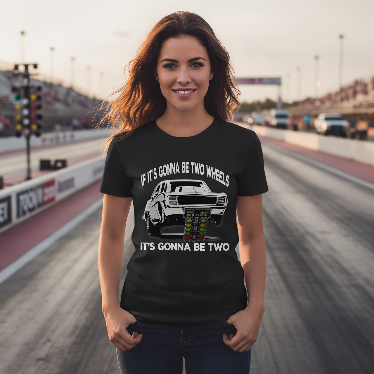 A woman stands on a racetrack wearing a black graphic T-shirt with a white car print and the text 'IF IT'S GONNA BE TWO WHEELS IT'S GONNA BE TWO'.