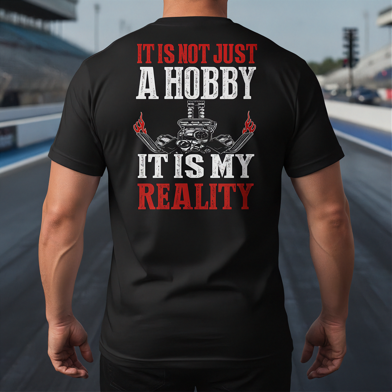A man viewed from behind wearing a black t-shirt with a large graphic print on the back. The graphic features the text 'IT IS NOT JUST A HOBBY IT IS MY REALITY' in white and red lettering, along with a black and red engine illustration.