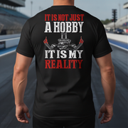 A man viewed from behind wearing a black t-shirt with a large graphic print on the back. The graphic features the text 'IT IS NOT JUST A HOBBY IT IS MY REALITY' in white and red lettering, along with a black and red engine illustration.