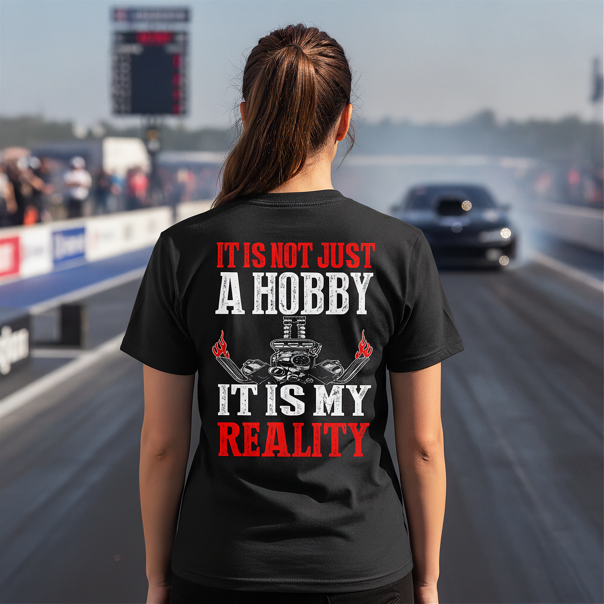 A woman stands on a racetrack from behind wearing a black graphic t-shirt with the text 'IT IS NOT JUST A HOBBY IT IS MY REALITY' in red and white, with a small engine graphic in the center. A drag race car is visible in the blurred background.