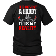 A black short-sleeve t-shirt shown from the back, featuring a bold graphic print with the text 'IT IS NOT JUST A HOBBY IT IS MY REALITY' in red and white, flanking a white skull and grill graphic with red flames.