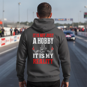 A man viewed from behind wearing a dark gray hoodie with a large graphic print on the back. The print features the text 'IT IS NOT JUST A HOBBY IT IS MY REALITY' in red and white, along with a small drag racing engine graphic.