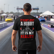 A man viewed from behind wearing a black graphic t-shirt with the text 'IT IS NOT JUST A HOBBY IT IS MY REALITY' in red and white, standing on a racetrack with cars blurred in the background.