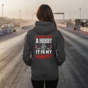 A woman stands on a racetrack wearing a dark gray hoodie from the back, which features a graphic print with the text 'IT IS NOT JUST A HOBBY IT IS MY REALITY' in red and white alongside a small illustration of a race car and checkered flag.