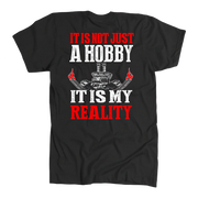 A black short-sleeve t-shirt with a graphic print on the front. The design features the text 'IT IS NOT JUST A HOBBY IT IS MY REALITY' in distressed white and red lettering, with a central illustration of a skull wearing a chef's hat and crossed tools, flanked by two hands holding red flags.