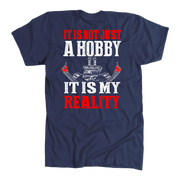 A navy blue short-sleeve t-shirt with a graphic print on the front. The design features the text 'IT IS NOT JUST A HOBBY IT IS MY REALITY' in distressed white and red lettering, with a central white skeleton key graphic and small red flame details, displayed on a clean white background.