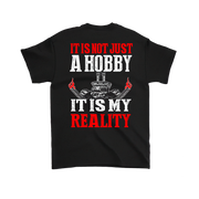 A black short-sleeve t-shirt shown from the back, featuring a bold graphic print with the text 'IT IS NOT JUST A HOBBY IT IS MY REALITY' in red and white distressed lettering, flanking a skull and crossed tools graphic.