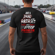 A man viewed from behind wearing a black t-shirt with a graphic on the back that reads 'JUST A DRAG RACER WITH A SPEED PROBLEM' in white and red lettering, set against a blurred drag racing track background.