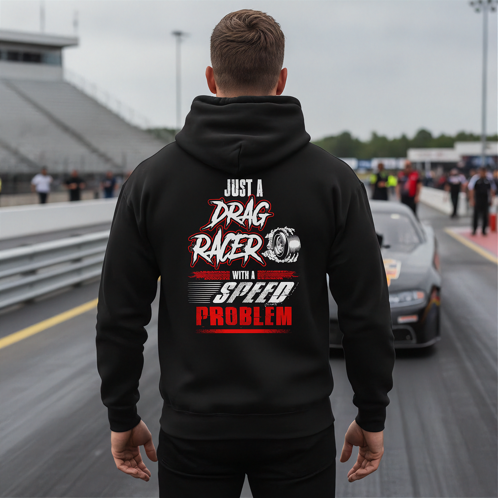 A man viewed from behind wearing a black hoodie with a large drag racing graphic on the back, standing on a racetrack with a car and grandstands in the background.