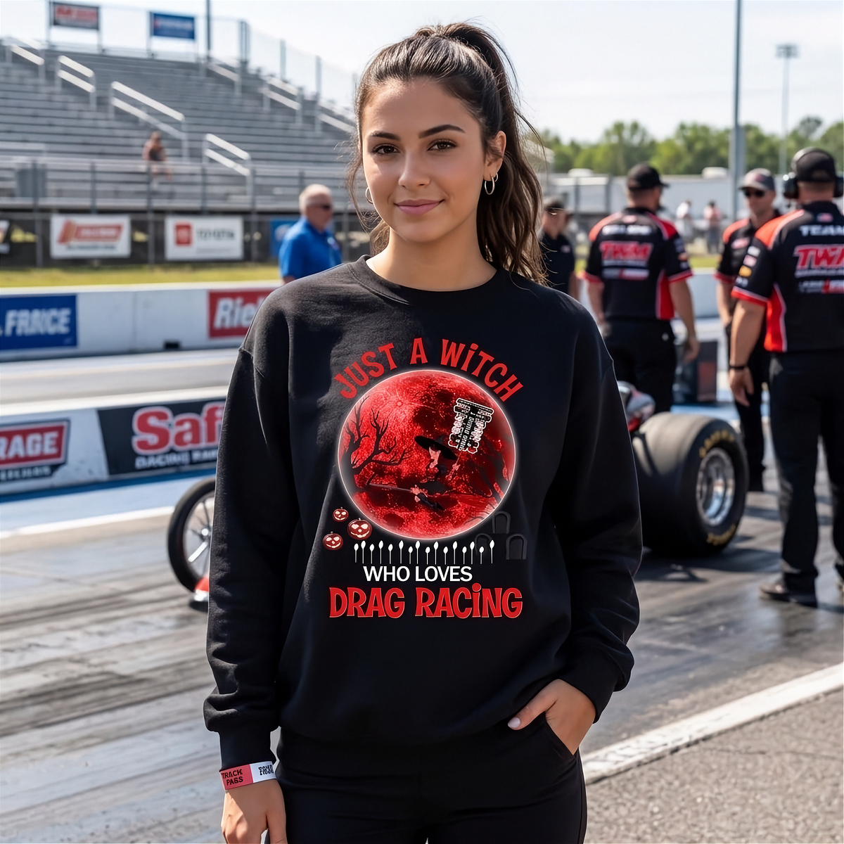 A woman stands on a drag racing track wearing a black crewneck sweatshirt with a red and black graphic of a witch riding a horse through a race track, with the text 'JUST A WITCH WHO LOVES DRAG RACING'.