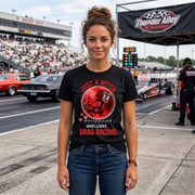 A woman stands on a racetrack wearing a black graphic T-shirt with a red circular design and the text 'Just a witch who loves drag racing' in red. Drag racing cars and a grandstands crowd are visible in the background under a Thunder Alley Racing banner.