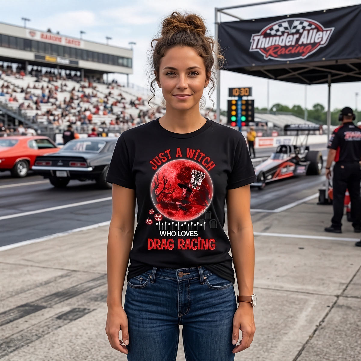 A woman stands on a racetrack wearing a black graphic T-shirt with a red circular design and the text 'Just a witch who loves drag racing' in red. Drag racing cars and a grandstands crowd are visible in the background under a Thunder Alley Racing banner.