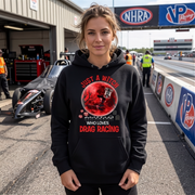 A woman stands on a drag racing track wearing a black hoodie with a red and white graphic that reads 'JUST A WITCH WHO LOVES DRAG RACING'. The background shows a race track pit area with a drag car, crew members, and NHRA and VP Racing banners.
