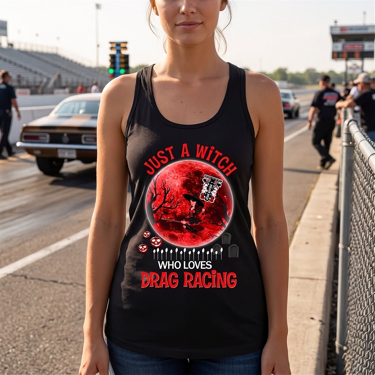 A woman wearing a black racerback tank top with a red and white graphic design that reads 'JUST A WITCH WHO LOVES DRAG RACING' stands on a racetrack. The graphic features a red full moon, a gothic cross, and small skulls.