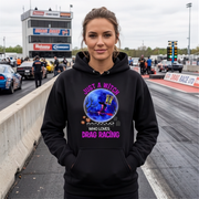 A woman stands on a drag racing track wearing a black hoodie with a colorful graphic of a witch riding a broom cart and the text 'JUST A WITCH WHO LOVES DRAG RACING' in pink and purple.