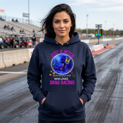A woman stands on a drag racing track wearing a navy blue hoodie with a colorful graphic that reads 'JUST A WITCH WHO LOVES DRAG RACING' around a witch and pumpkin illustration.