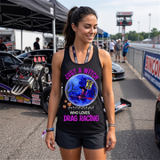 A woman stands in a drag racing pit area wearing a black tank top with a colorful graphic of a witch riding a broomstick on a blue moon, surrounded by pumpkins, with the text 'JUST A WITCH WHO LOVES DRAG RACING'.