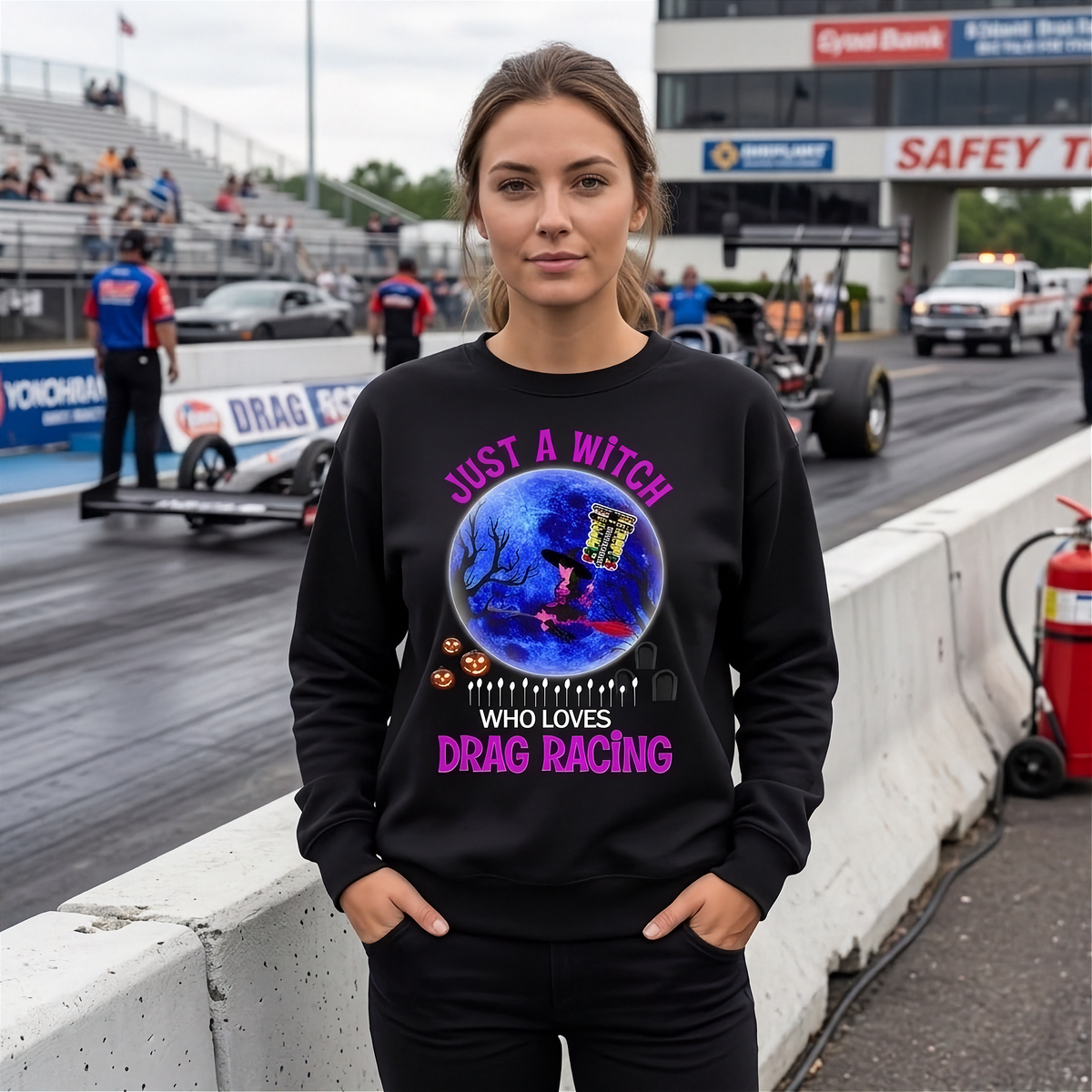 A woman stands at a drag racing track wearing a black crewneck sweatshirt with a colorful graphic on the chest. The graphic features a blue and purple moon with a witch on a broomstick, surrounded by the text 'JUST A WITCH WHO LOVES DRAG RACING' in pink and white.