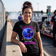 A woman stands on a racetrack wearing a black graphic T-shirt with a Halloween-themed drag racing design. The shirt features the text 'JUST A WITCH WHO LOVES DRAG RACING' in pink and white, surrounding a circular blue and purple illustration of a witch riding a broomstick in a starry night sky.