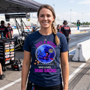 A smiling woman stands on a drag racing track wearing a navy blue graphic t-shirt with a witch and drag racing theme. The background shows a pit lane environment with race equipment, crew, and track barriers.