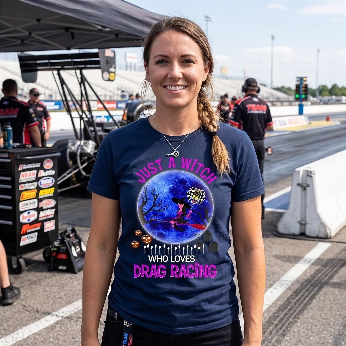 A smiling woman stands on a drag racing track wearing a navy blue graphic t-shirt with a witch and drag racing theme. The background shows a pit lane environment with race equipment, crew, and track barriers.