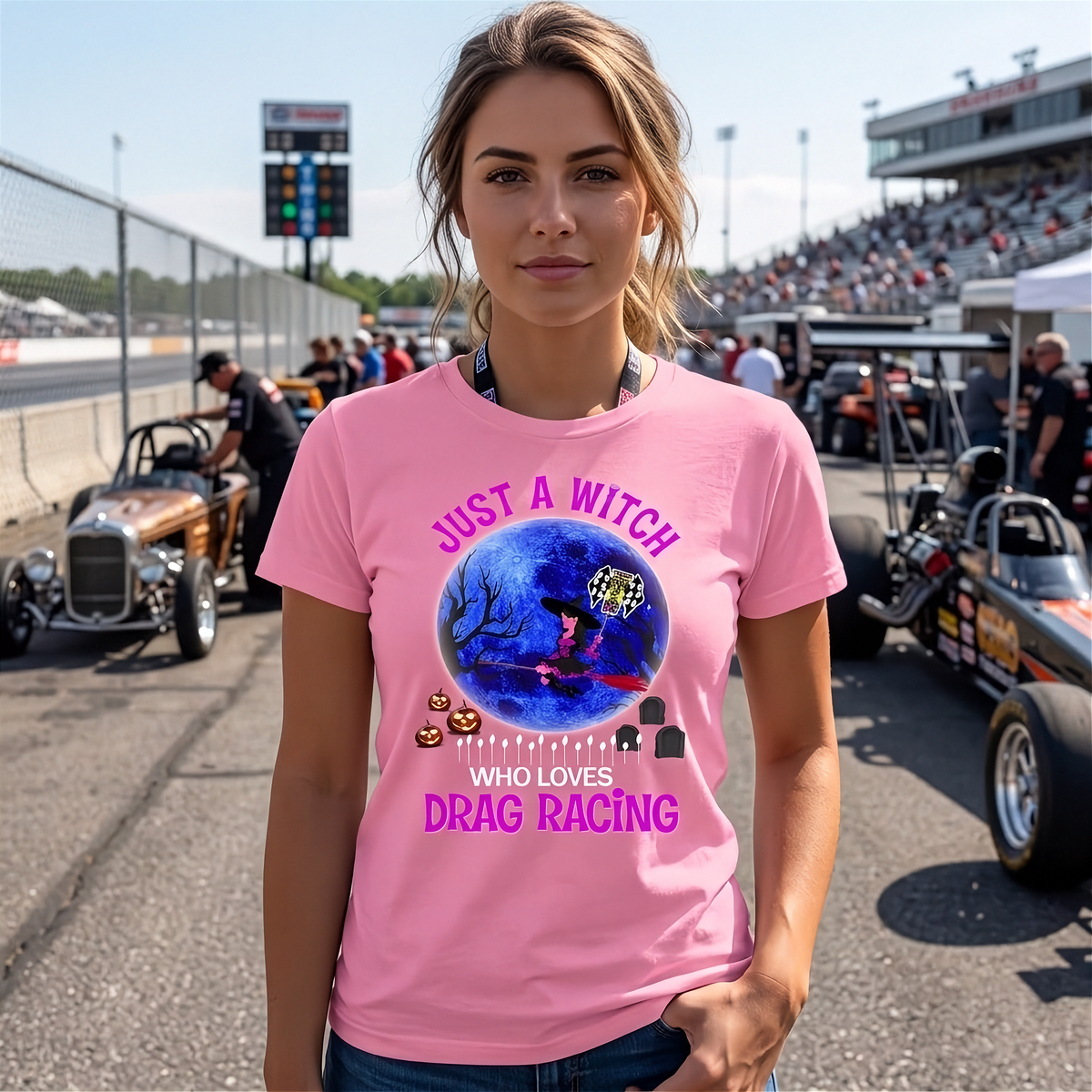 A woman wearing a pink graphic t-shirt with a Halloween-themed drag racing design, standing on a racetrack with race cars and grandstands in the background.