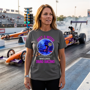 A woman stands on a drag racing track wearing a dark gray graphic t-shirt with a Halloween-themed design featuring a witch, pumpkin, and the text 'JUST A WITCH WHO LOVES DRAG RACING' in pink. Drag races and racecars are visible in the background.
