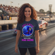 A woman with curly brown hair stands on a drag racing track wearing a navy blue graphic t-shirt. The shirt features a circular illustration of a witch riding a broom cart in a blue moonlit sky, surrounded by the text 'JUST A WITCH WHO LOVES DRAG RACING' in pink and white.