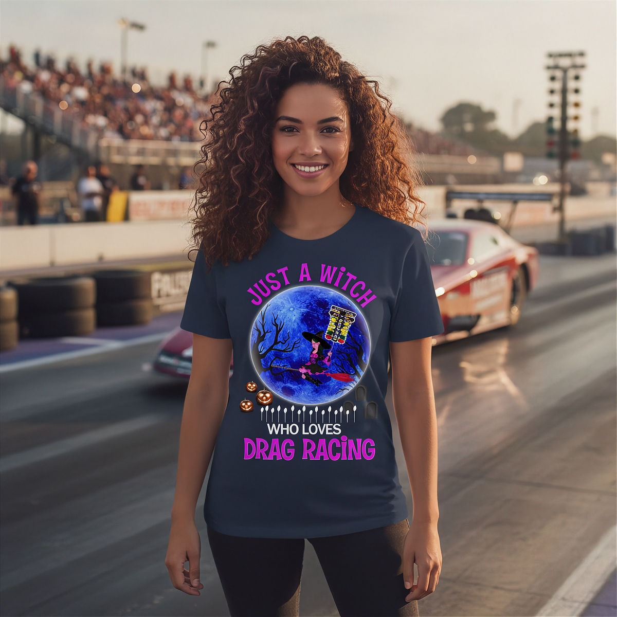 A woman with curly brown hair stands on a drag racing track wearing a navy blue graphic t-shirt. The shirt features a circular illustration of a witch riding a broom cart in a blue moonlit sky, surrounded by the text 'JUST A WITCH WHO LOVES DRAG RACING' in pink and white.