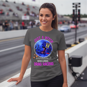 A woman wearing a dark gray graphic t-shirt with a Halloween-themed drag racing design, standing on a bridge with a blurred racetrack and grandstands in the background.