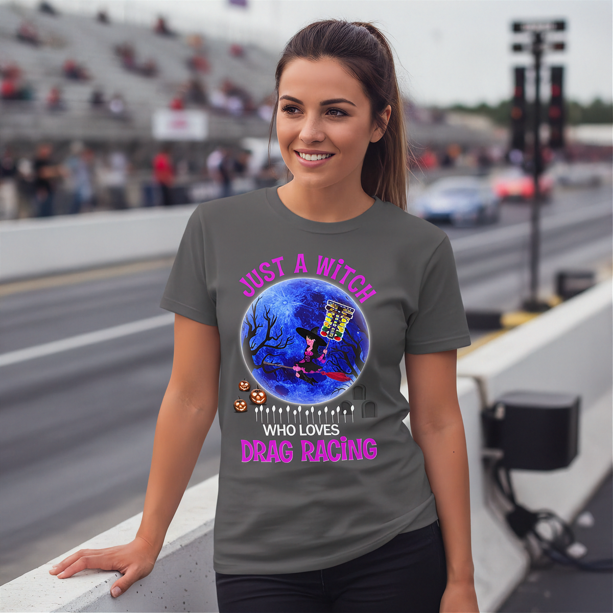 A woman wearing a dark gray graphic t-shirt with a Halloween-themed drag racing design, standing on a bridge with a blurred racetrack and grandstands in the background.