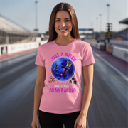 A woman stands on a racetrack wearing a light pink graphic T-shirt with a Halloween-themed illustration and the text 'JUST A WITCH WHO LOVES DRAG RACING'.