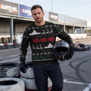 A man wearing a black Christmas sweater with red 'HO-HO-HO' text and white holiday graphics stands on a racetrack holding a black helmet. Karts Inc.