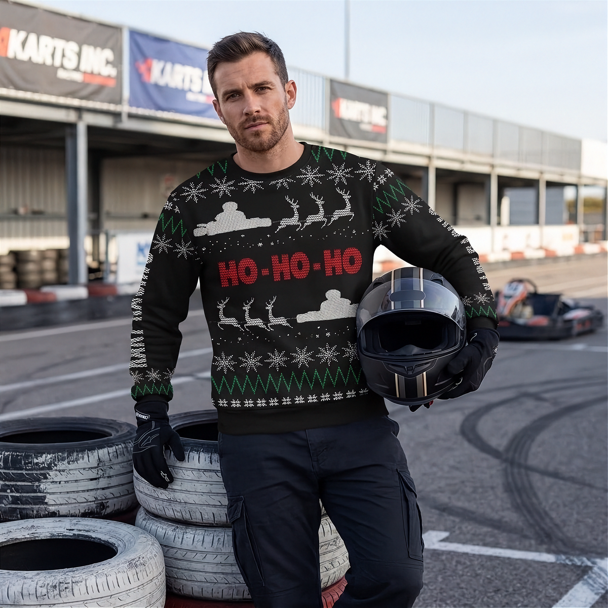 A man wearing a black Christmas sweater with red 'HO-HO-HO' text and white holiday graphics stands on a racetrack holding a black helmet. Karts Inc.