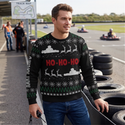 A man wearing a black Christmas sweater with a festive holiday pattern and 'HO-HO-HO' text in red, standing on a race track with go-karts in the background.