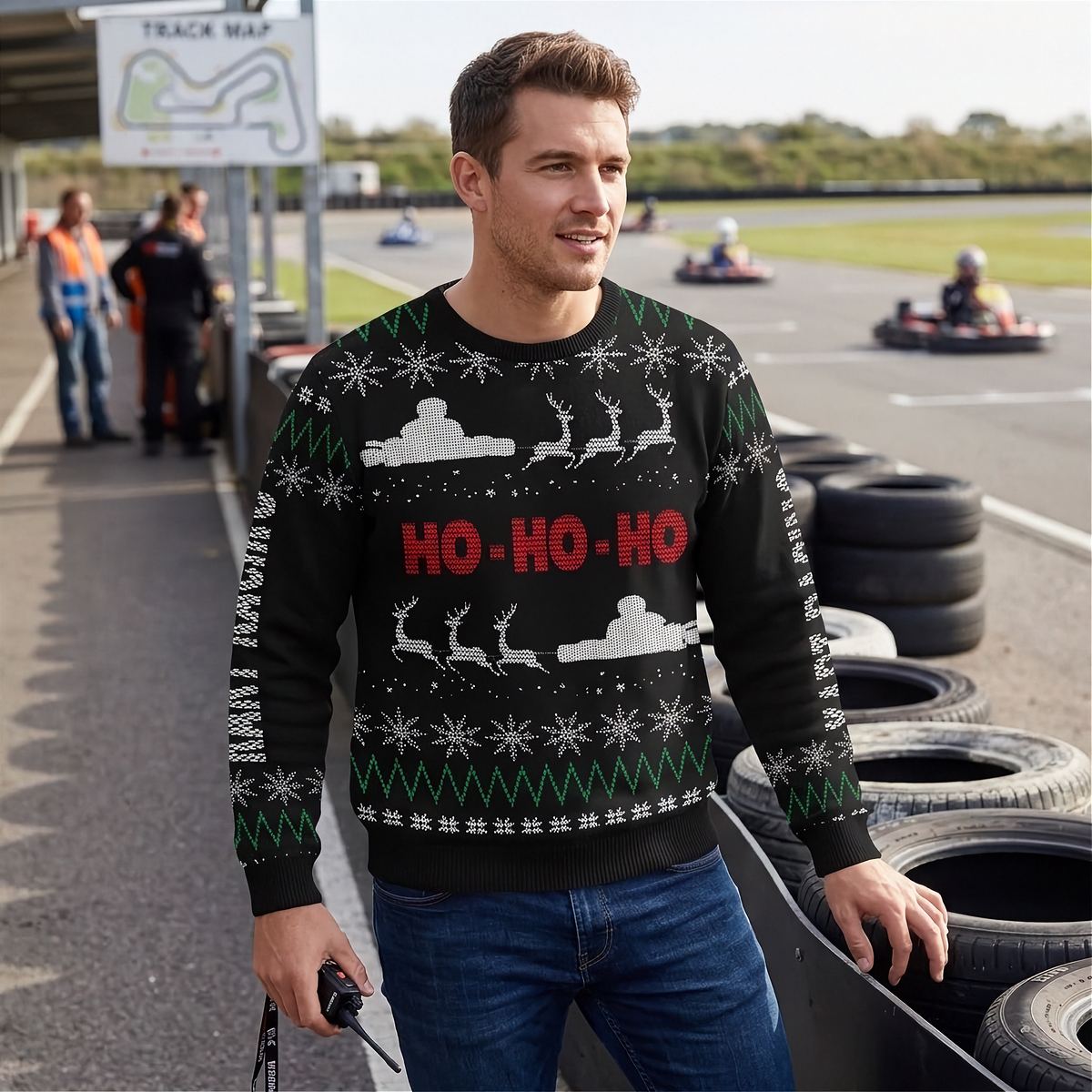 A man wearing a black Christmas sweater with a festive holiday pattern and 'HO-HO-HO' text in red, standing on a race track with go-karts in the background.