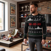 A man wearing a black Christmas sweater with a white Santa, reindeer, snowflakes, and red 'HO-HO-HO' text, standing in a modern room with go-karts and racing posters in the background.