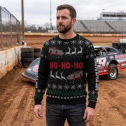 A man wearing a black Christmas sweater with a red and gray race car and sledding pattern stands on a dirt racetrack with race cars and grandstands in the background.