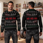 A man modeling a black Christmas sweater in a room, shown from both the front and back. The sweater features a festive holiday pattern with red 'HO-HO-HO' text, white reindeer, and red-and-white trucks, accented with green and white snowflake motifs.