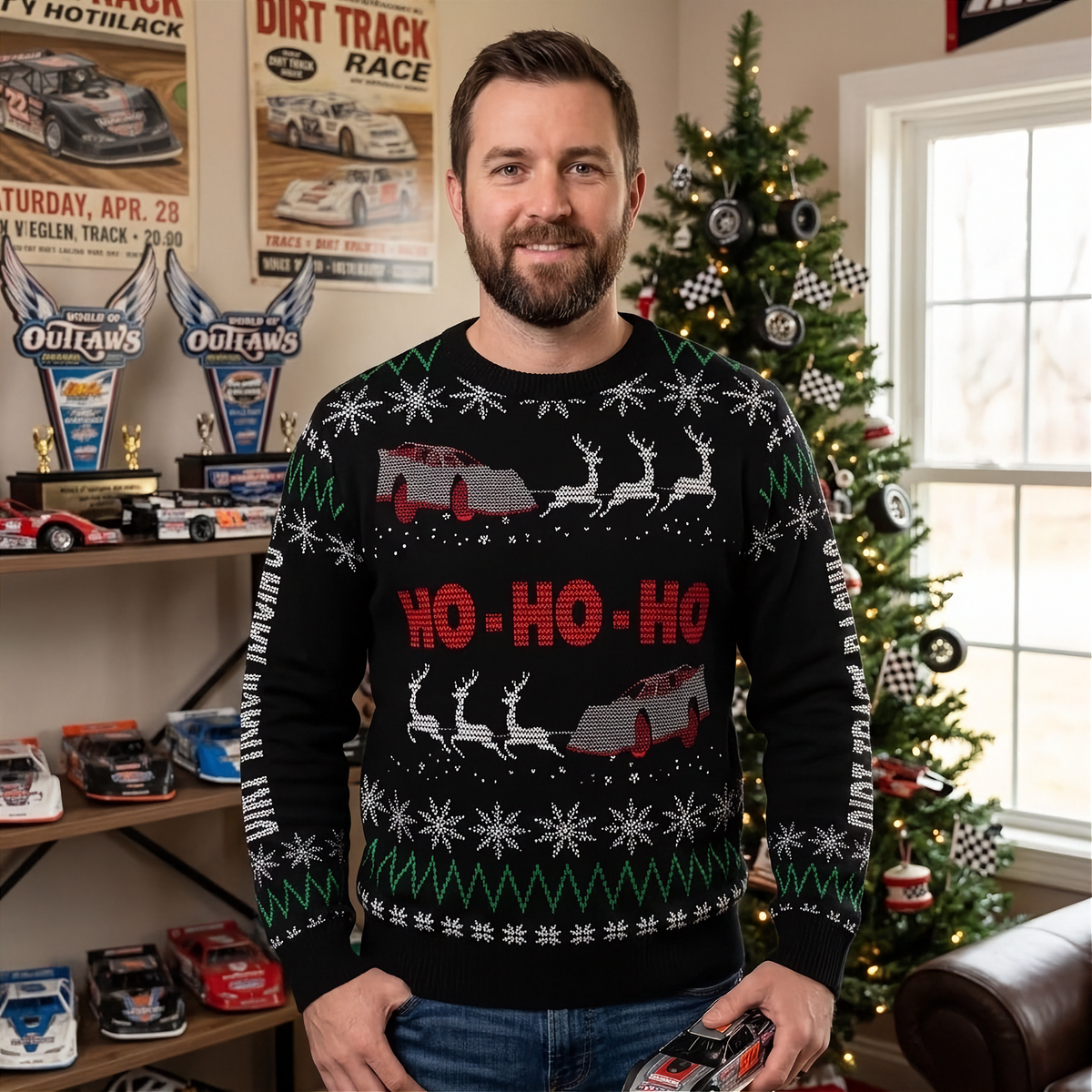 A man wearing a black Christmas sweater with a holiday racing theme, standing in a decorated room with a Christmas tree and racing memorabilia in the background.