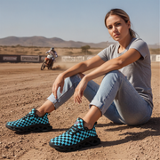 Woman sitting on a dirt track wearing blue checkered shoes with a motorcycle in the background.