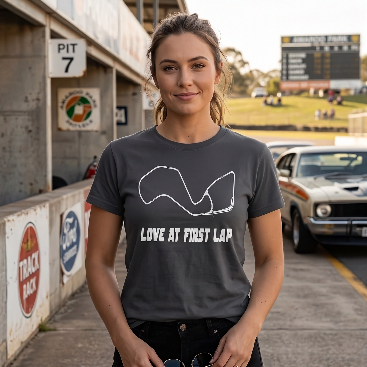A woman stands on a racetrack wearing a dark gray graphic t-shirt with a white track outline and the text 'LOVE AT FIRST LAP'. Classic cars and track signage are visible in the background.