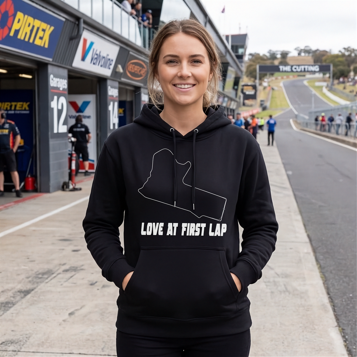 A woman stands on a race track pit lane wearing a black hoodie with a white outlined track map and the text 'LOVE AT FIRST LAP' on the front.