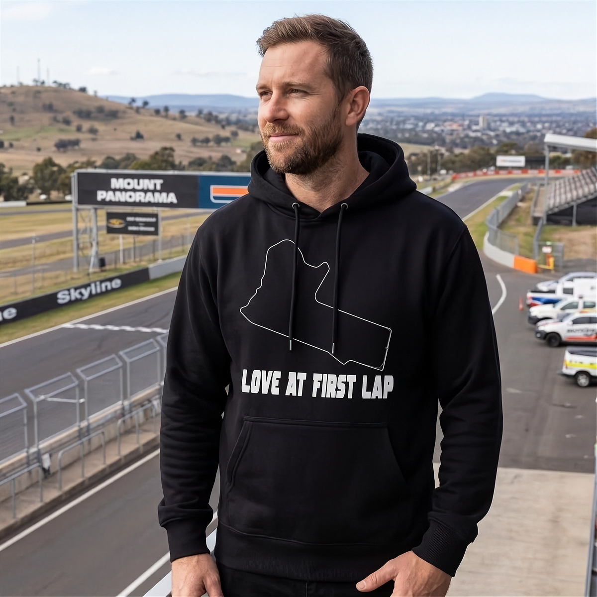 A man wearing a black pullover hoodie with a white graphic of the Australian map outline and the text 'LOVE AT FIRST LAP' stands on a race track overlooking a circuit.