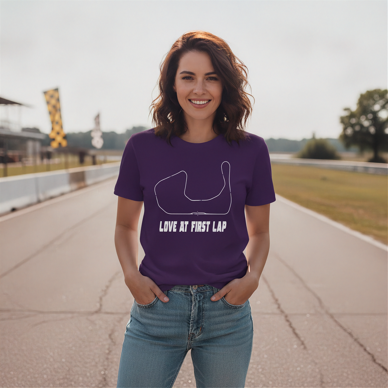 A woman stands on a racetrack wearing a purple short-sleeve graphic T-shirt with a white track outline and the text 'LOVE AT FIRST LAP,' paired with light blue jeans.