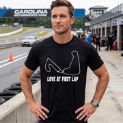 A man wearing a black t-shirt with a white graphic of a racing arm and the text 'LOVE AT FIRST LAP' stands on a racetrack. The background shows the track, pit stops, and a sign for Carolina Motorsports Park.