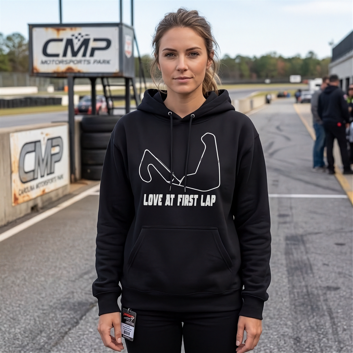A woman stands on a racetrack wearing a black pullover hoodie with a white graphic of a flexed arm and the text 'LOVE AT FIRST LAP'. The background shows CMP Motorsports Park signage and the track surface.