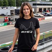 A woman stands on a racetrack wearing a black graphic T-shirt with a white circuit-racing-style line drawing and the text 'LOVE AT FIRST LAP.' The background shows race track equipment, trailers, cars, fencing, and a building labeled Carolina Motorsports Park.